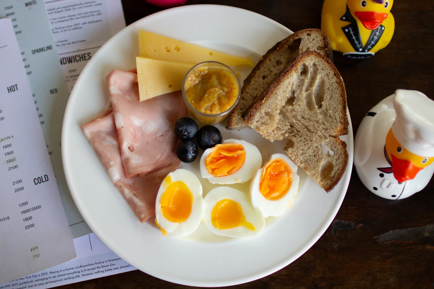 Breakfast plate with soft-boiled eggs, cheese, mortadella, bread, olives, and jam